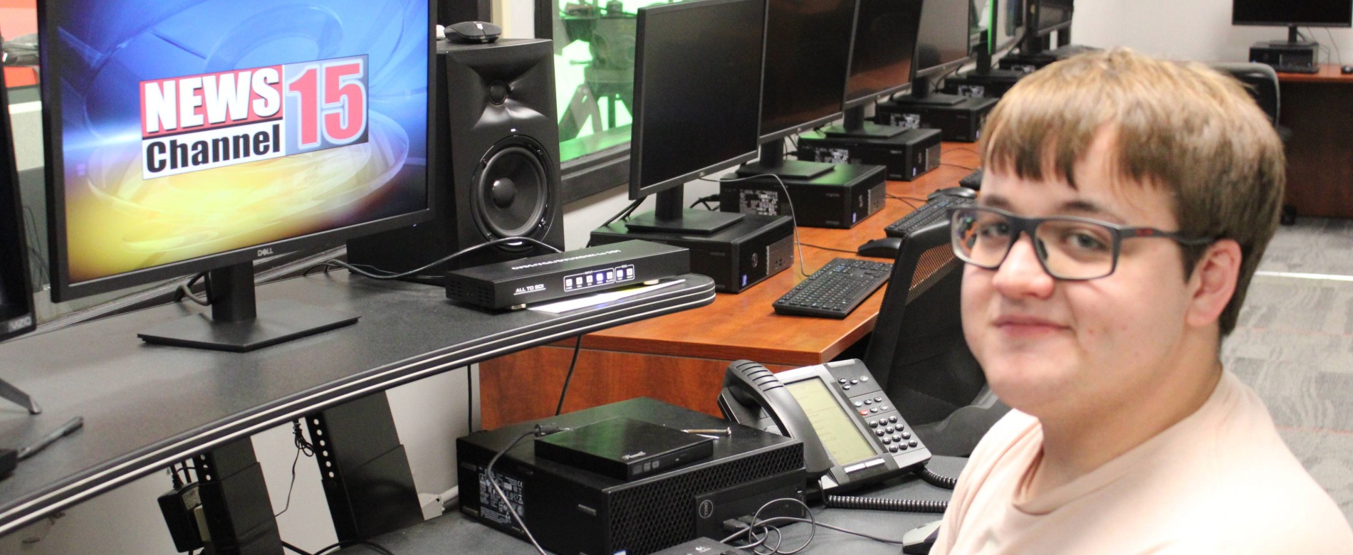 WVC student seated at a workstation with multiple computer monitors and broadcast equipment. One monitor displays a screen with the text ‘NEWS Channel 15.’ The desk includes a keyboard, control panel, phone, and audio equipment.