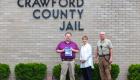 Fred Chinn, Rena Gower, Bill Rutan stand in front of Crawford County Jail holding donated book